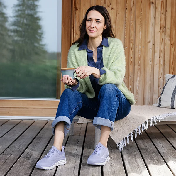 Lächelnde Dame mit Kaffee in der Hand sitzt auf einer Liege mit bequemen Sportschuhen.