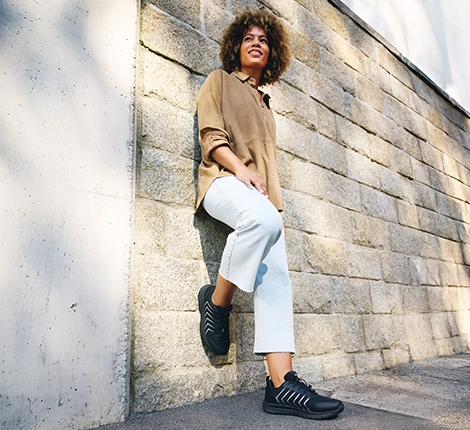 Junge Frau lehnt an einer Wand mit schwarzen Sportschuhen.